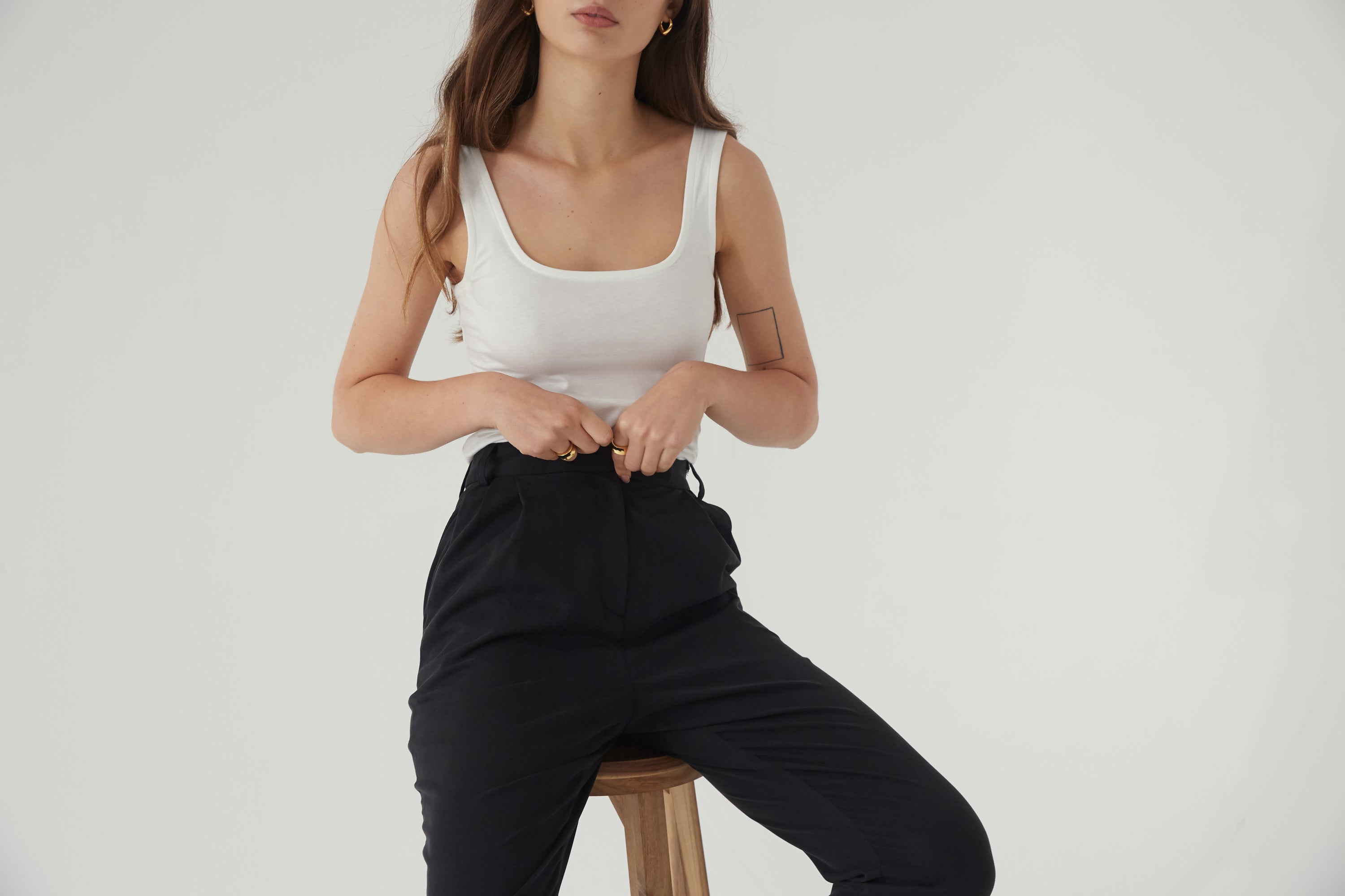 Woman wearing a white tank top and black pants sitting on a wooden stool against a plain background