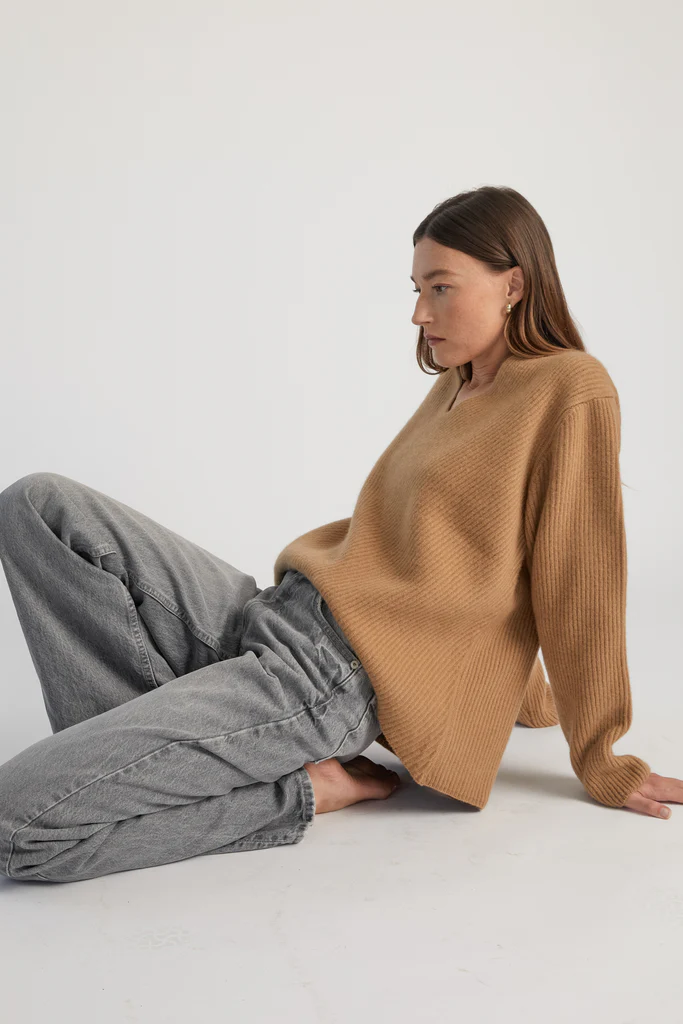 Woman wearing a brown sweater and gray pants sitting on a white floor.