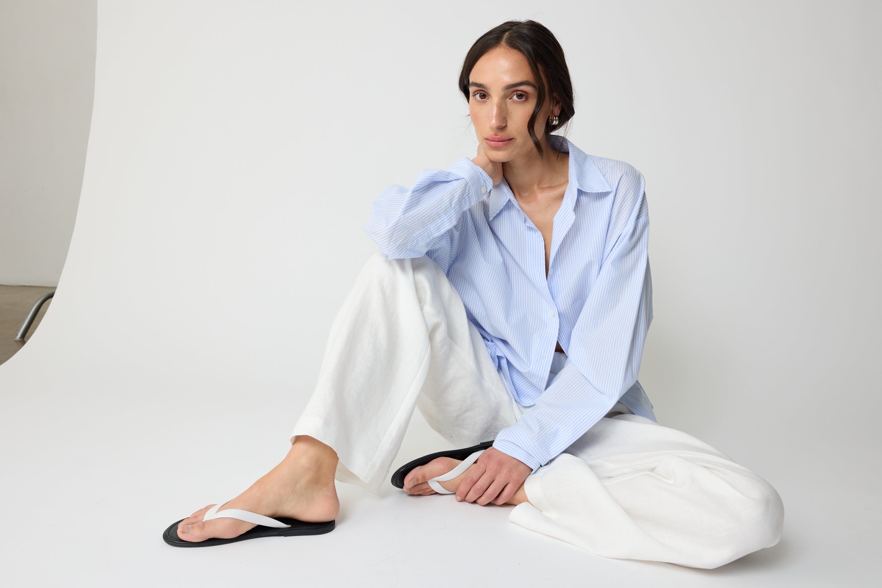 Woman wearing a light blue striped shirt and white pants sitting on a white surface.