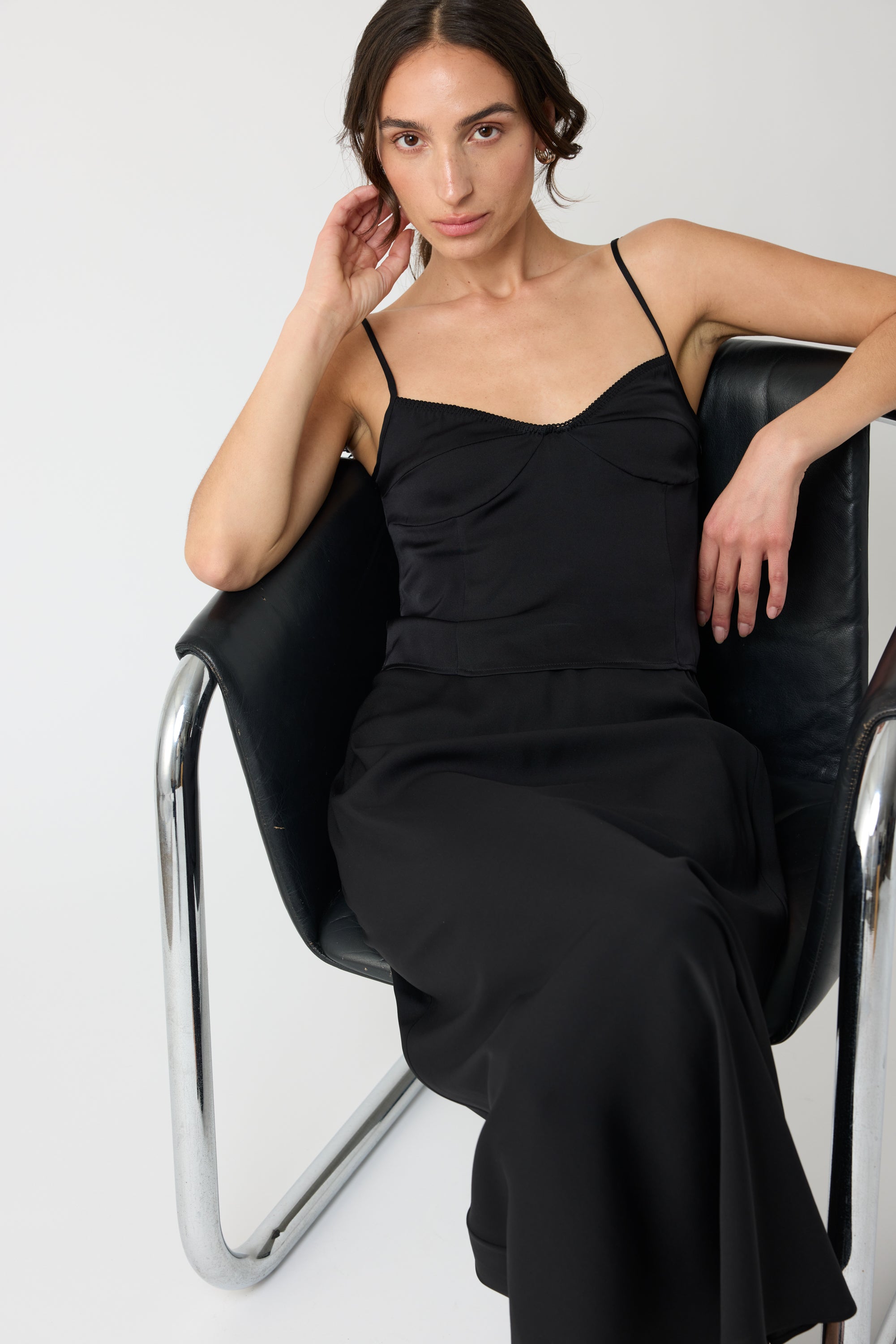 Woman wearing a black silk top and matching black silk skirt sitting on a chair against a white background