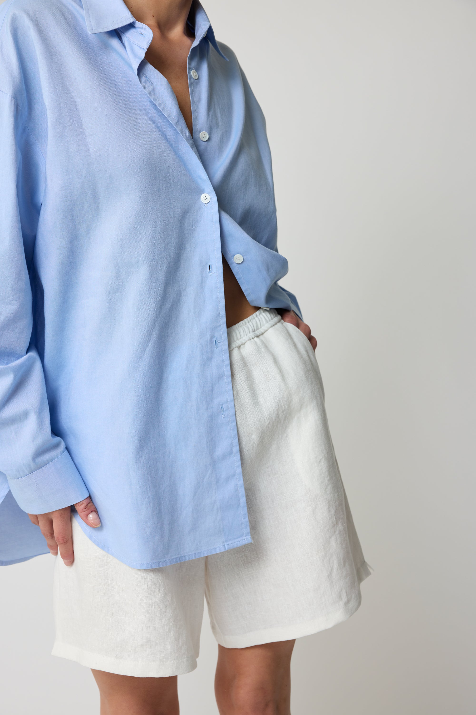 Woman wearing a light cotton button up blue shirt and white linen shorts on a plain background