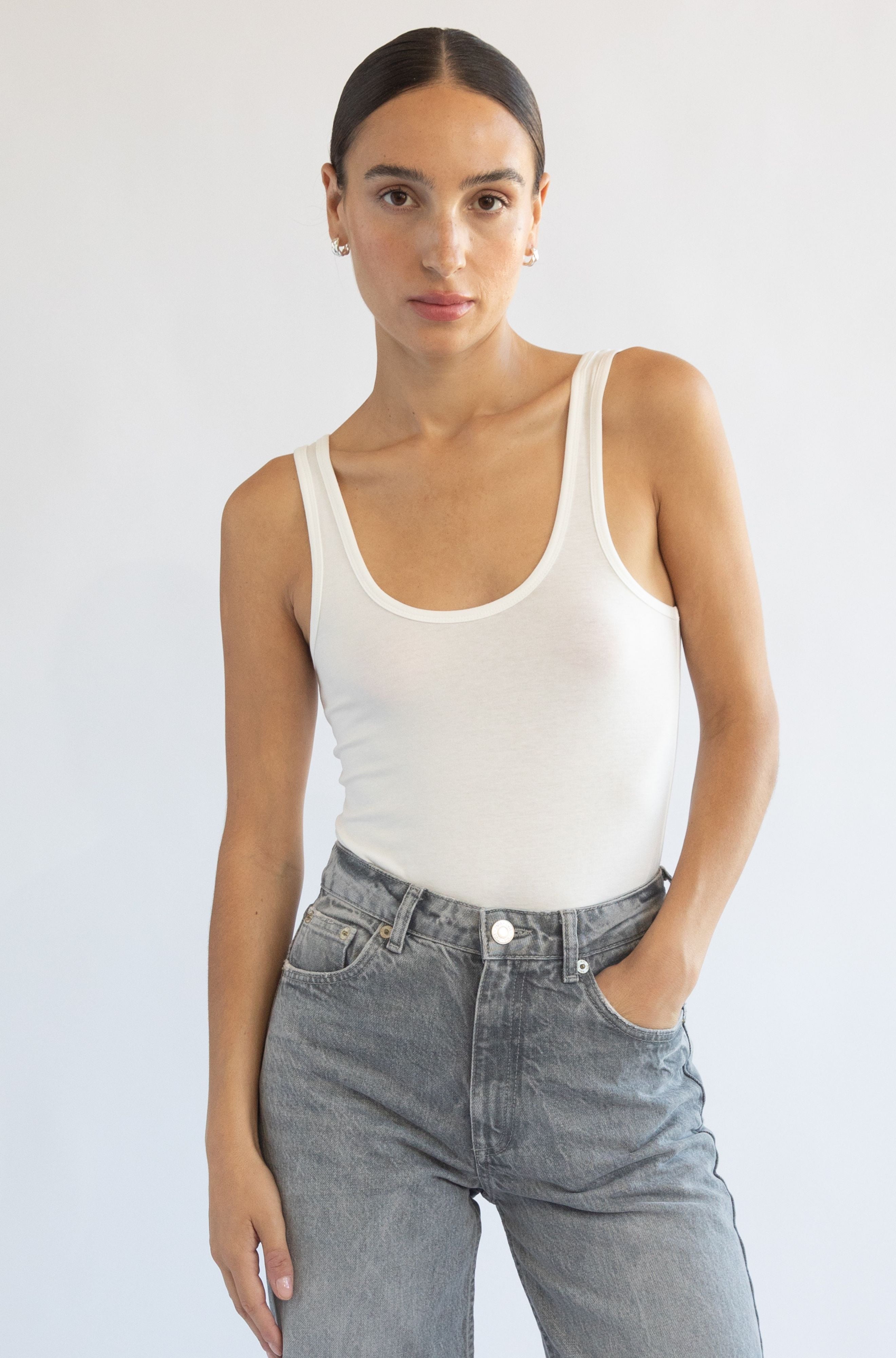 Woman wearing a white tank top and gray jeans against a plain background